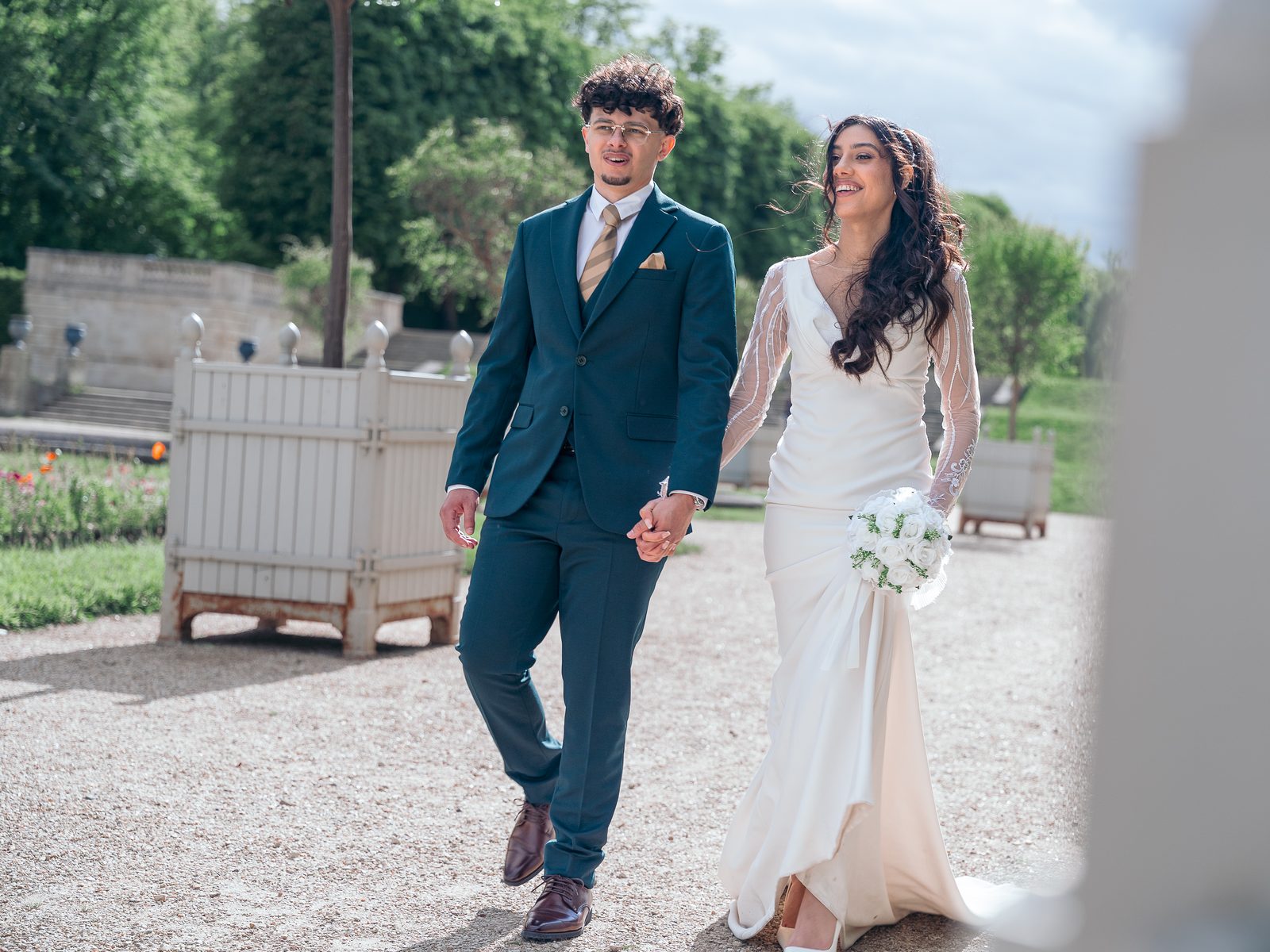 Les mariés avancent sur le gravier, la mariée éclate de rire, bouquet à la main